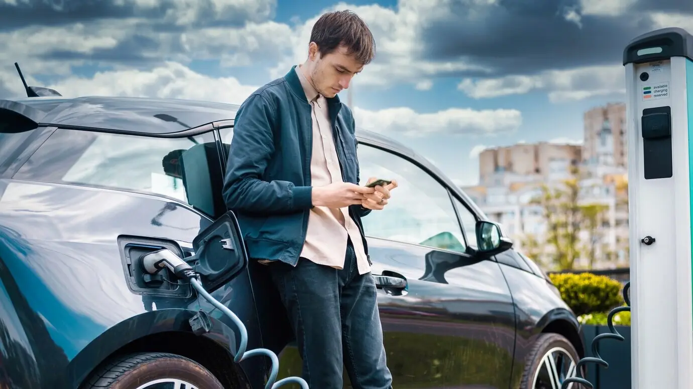 A man is charging his electric car at a charging station while using his smartphone.